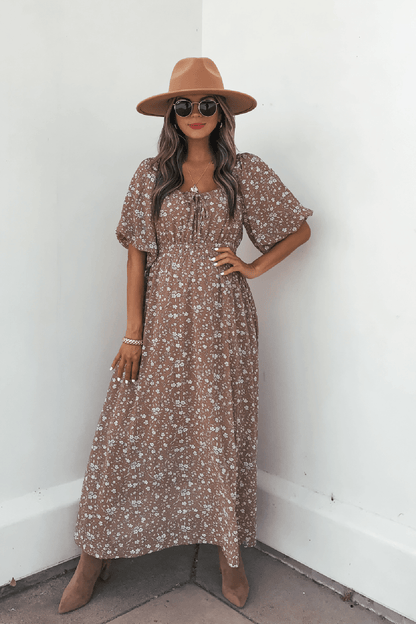Woman in Mocha Floral Print Smocked Midi Dress, tan hat, and sunglasses poses by a white wall with one hand on her hip.