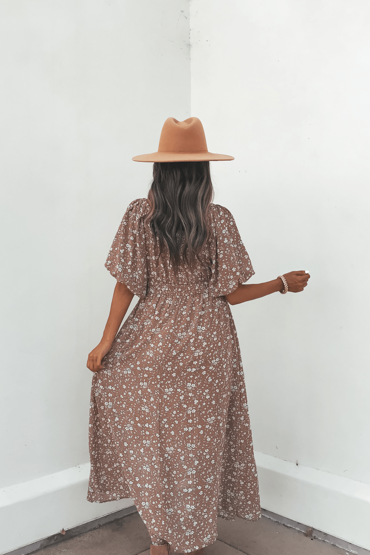 A woman in the Mocha Floral Print Smocked Midi Dress and a tan hat stands with her back to the camera against a white wall.