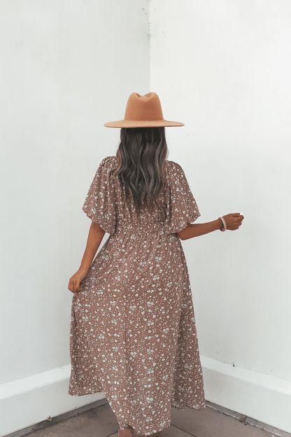 A woman in the Mocha Floral Print Smocked Midi Dress and a tan hat stands with her back to the camera against a white wall.