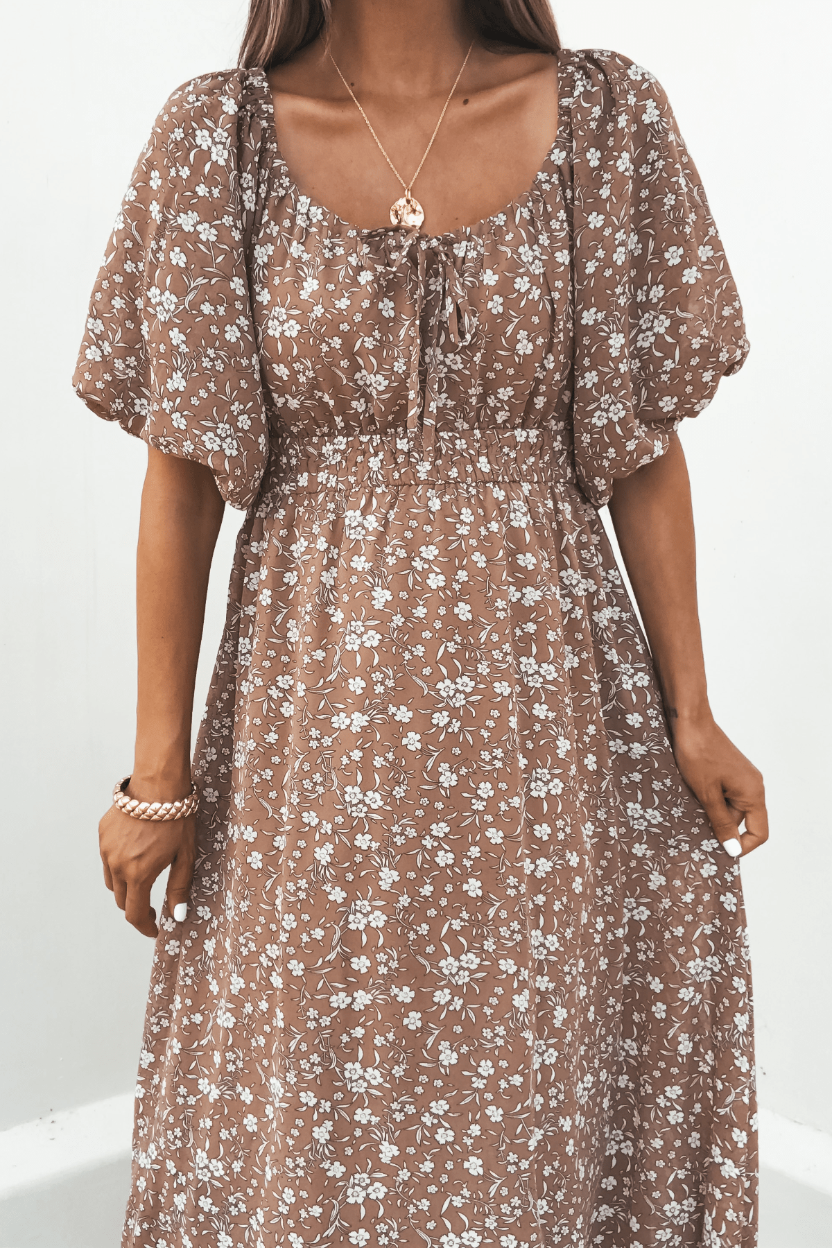A woman wears the Mocha Floral Print Smocked Midi Dress with puff sleeves and a gold necklace, standing against a white background.