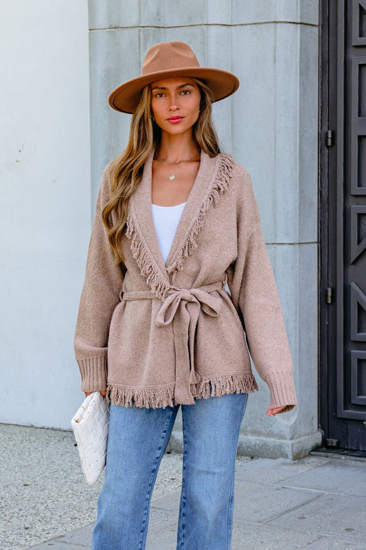 Woman in hat and Mocha Fringe Tie Cardigan stands outdoors holding a white clutch bag in front of a building.