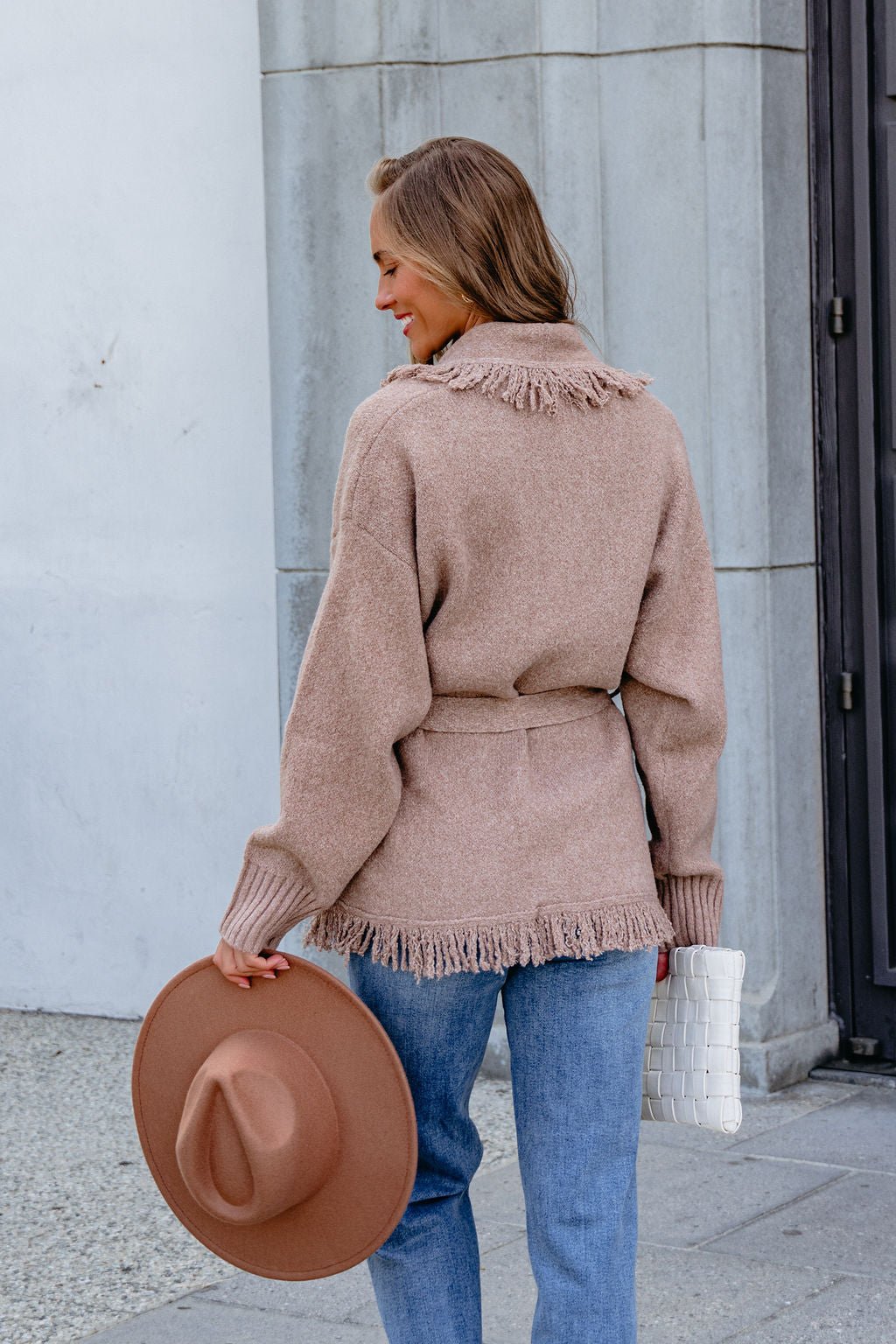 Woman in Mocha Fringe Tie Cardigan, jeans, holding a brown hat and white clutch, standing outdoors near a building.