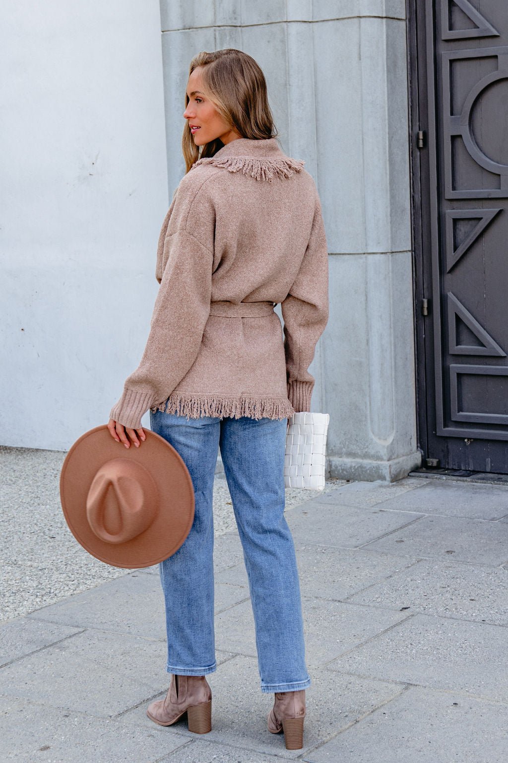 Woman in Mocha Fringe Tie Cardigan holds a brown hat and white bag near a gray stone building. Pre Order now.