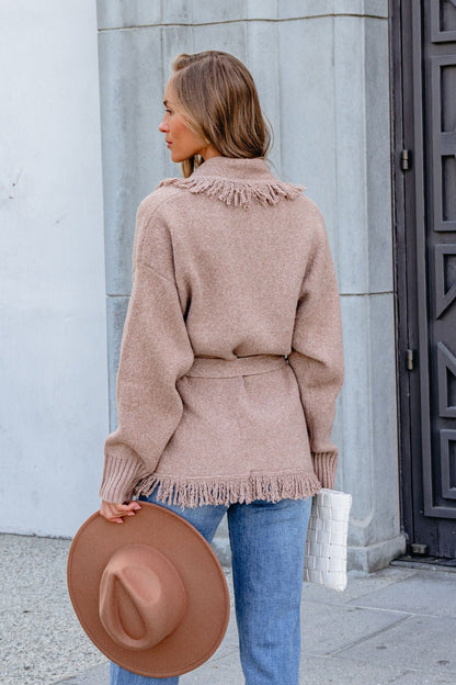 Woman stands outdoors in the Mocha Fringe Tie Cardigan, blue jeans, holding a tan hat and white clutch, facing away from the camera.