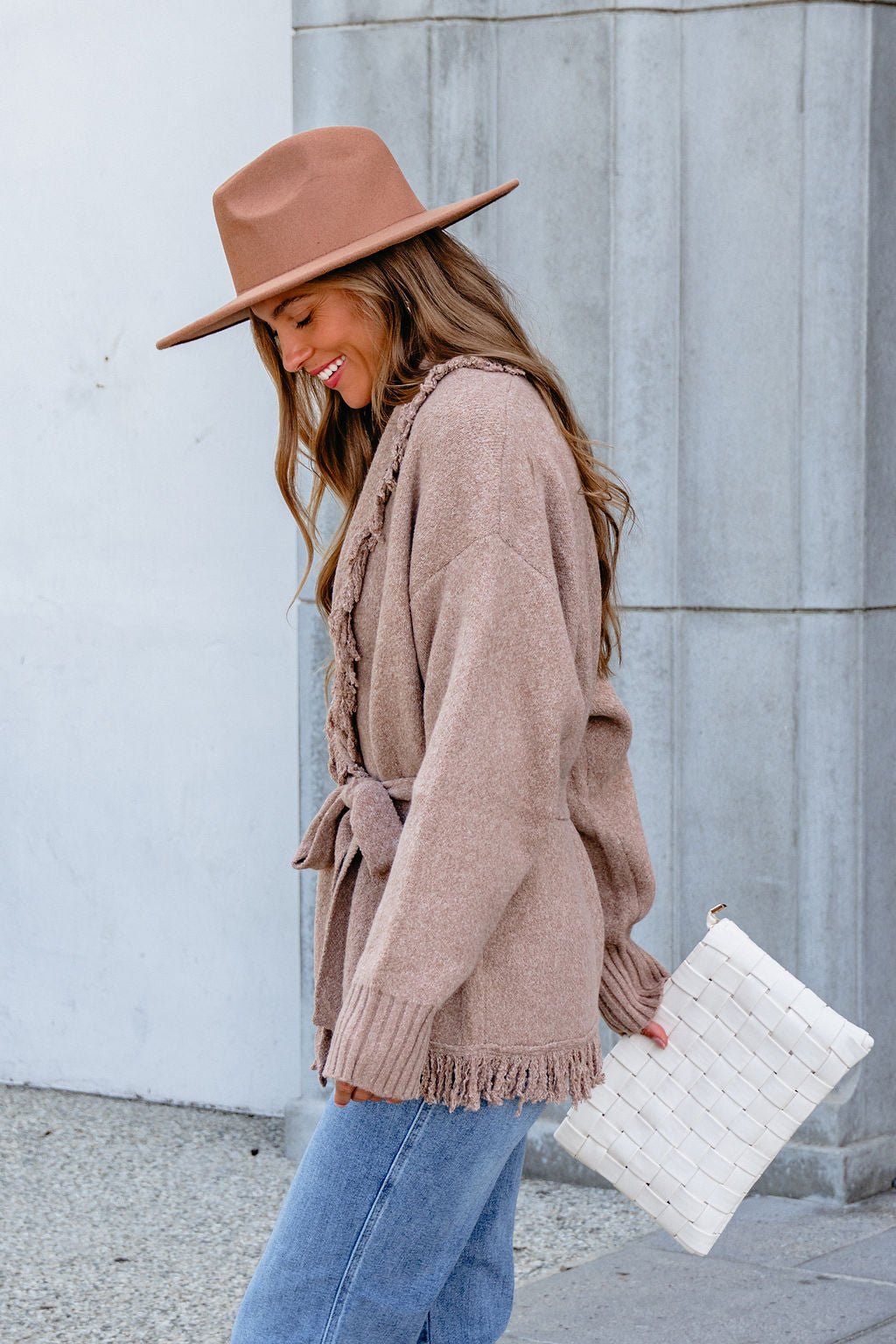 Smiling woman in a tan hat and blue jeans wears the Mocha Fringe Tie Cardigan (Pre Order) and holds a white woven clutch.