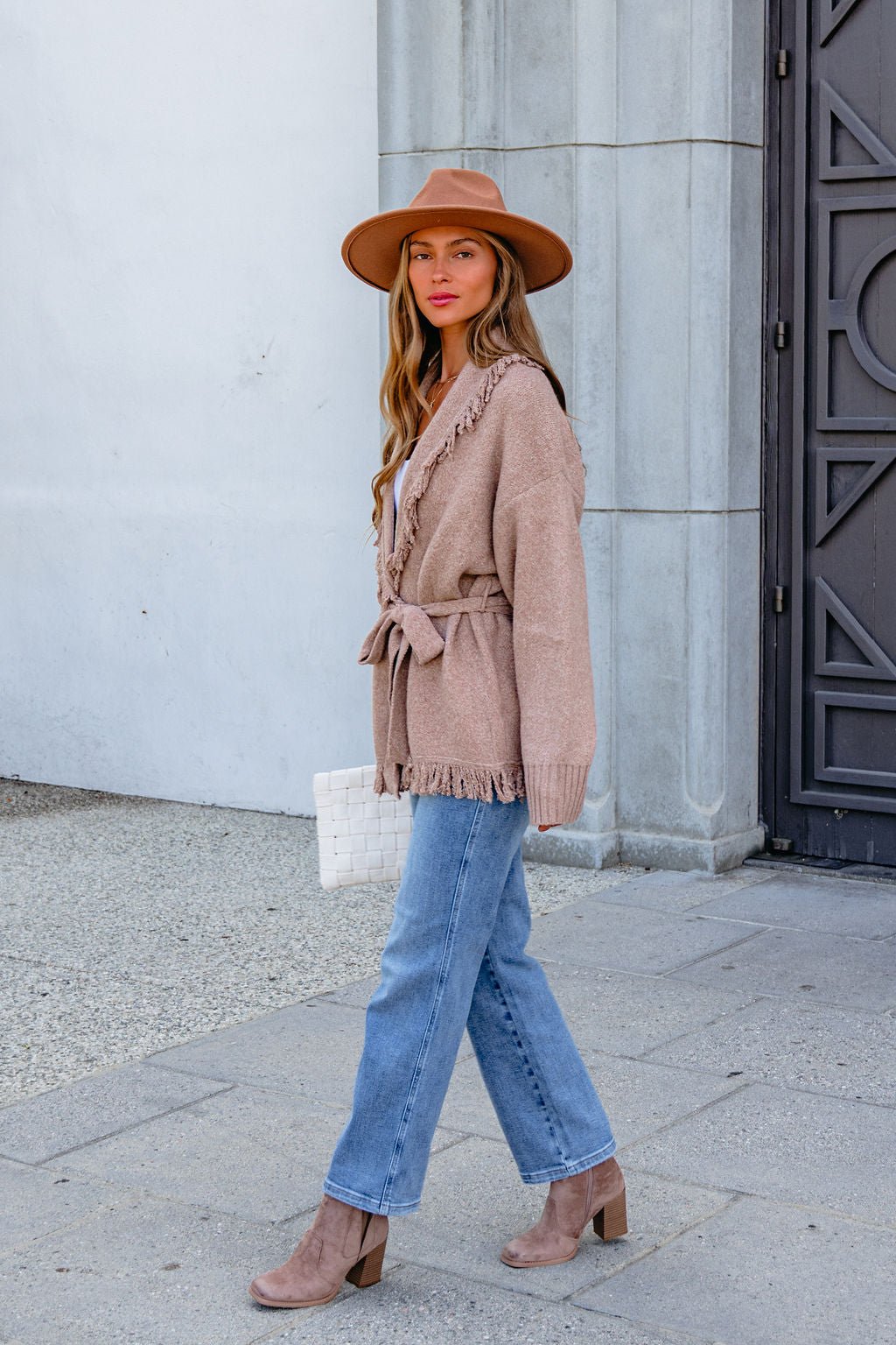 Woman in a tan hat, Mocha Fringe Tie Cardigan, blue jeans, and heeled boots walks holding a white woven bag.