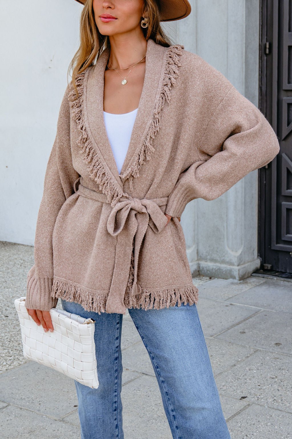 Woman in Mocha Fringe Tie Cardigan (preorder), blue jeans, white top, brown hat, holding a white woven clutch.