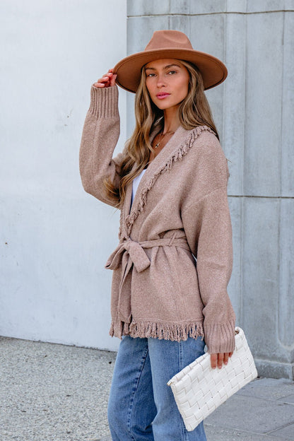 Woman in tan hat, jeans, and Mocha Fringe Tie Cardigan stands near a gray wall, holding a white woven clutch.