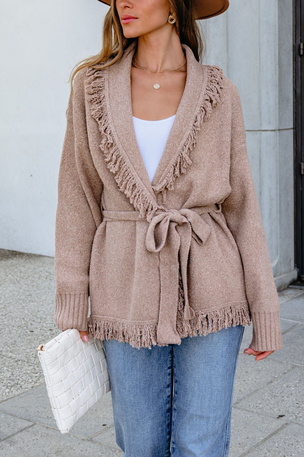 Woman in Mocha Fringe Tie Cardigan | Pre Order, white top, jeans, brown hat, holding a white clutch outside.