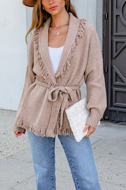 Woman in Mocha Fringe Tie Cardigan (Pre Order), blue jeans, and white woven clutch, standing outside.