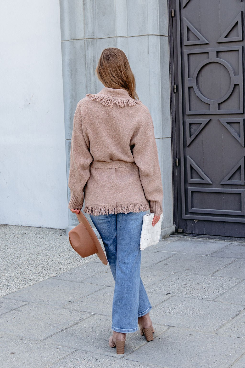 Person in Mocha Fringe Tie Cardigan, jeans, and heels stands outside facing away, holding a hat and white clutch bag.