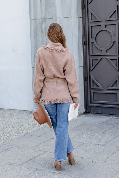 Person in Mocha Fringe Tie Cardigan, jeans, and heels stands outside facing away, holding a hat and white clutch bag.