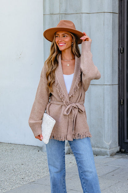 Woman in tan hat and Mocha Fringe Tie Cardigan (Pre Order) with jeans, white top, and clutch smiles outside a stone building.