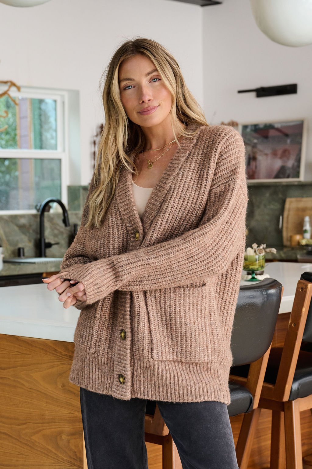 A woman wears the Mocha Heavy Knit Button Down Cardigan in a modern kitchen, smiling gently at the camera.