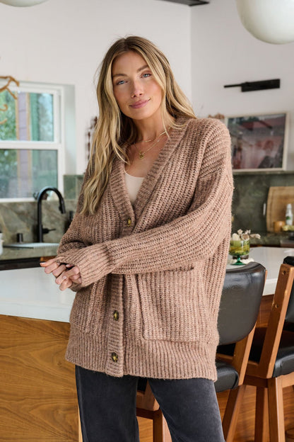 A woman wears the Mocha Heavy Knit Button Down Cardigan in a modern kitchen, smiling gently at the camera.