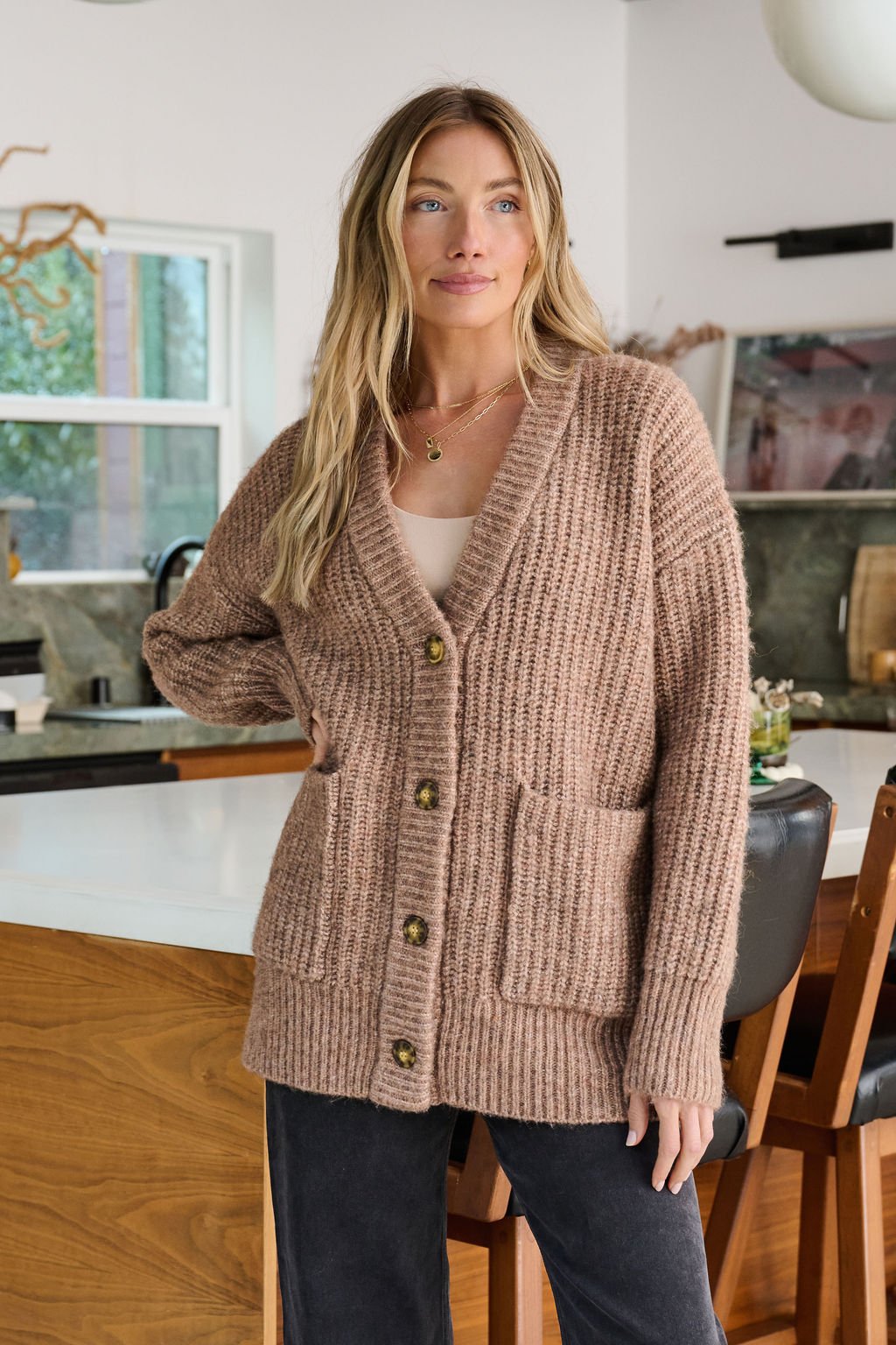 A woman stands in a modern kitchen wearing the Mocha Heavy Knit Button Down Cardigan with large buttons and front pockets.