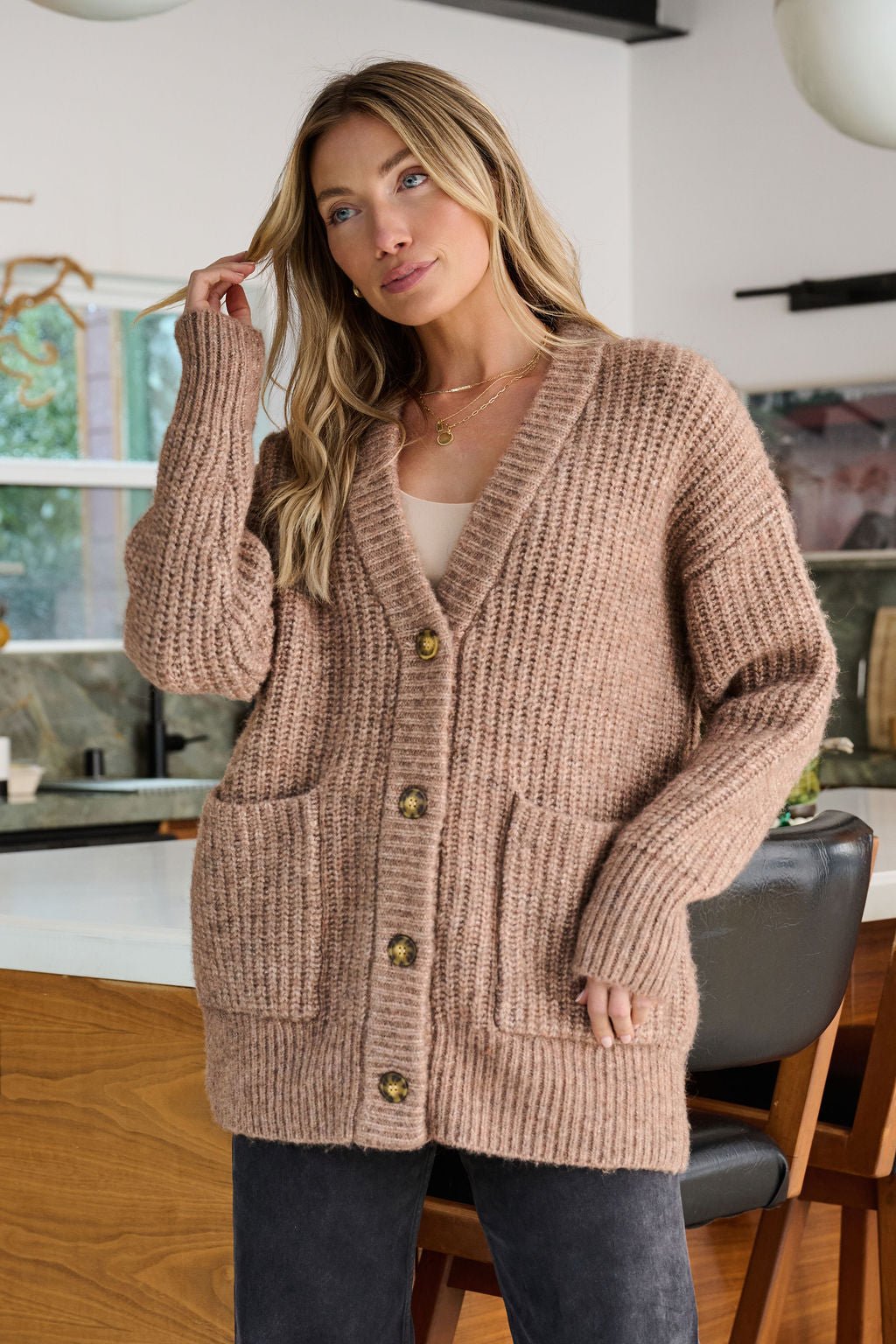 A woman stands indoors wearing the Mocha Heavy Knit Button Down Cardigan, featuring large front pockets and button details.