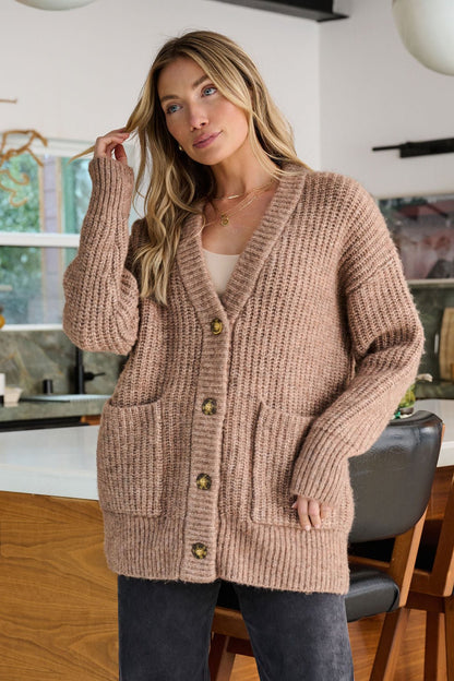 A woman stands indoors wearing the Mocha Heavy Knit Button Down Cardigan, featuring large front pockets and button details.