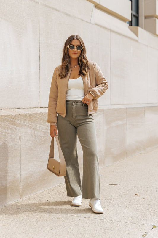 Woman in sunglasses, Tan Quilted Bomber Jacket, white top, gray pants, and sneakers stands on a sidewalk holding a beige handbag.