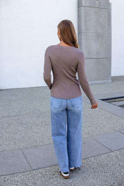 A person wears a Mocha Ribbed Peplum Sweater and blue jeans outdoors, long hair flowing as they face away.