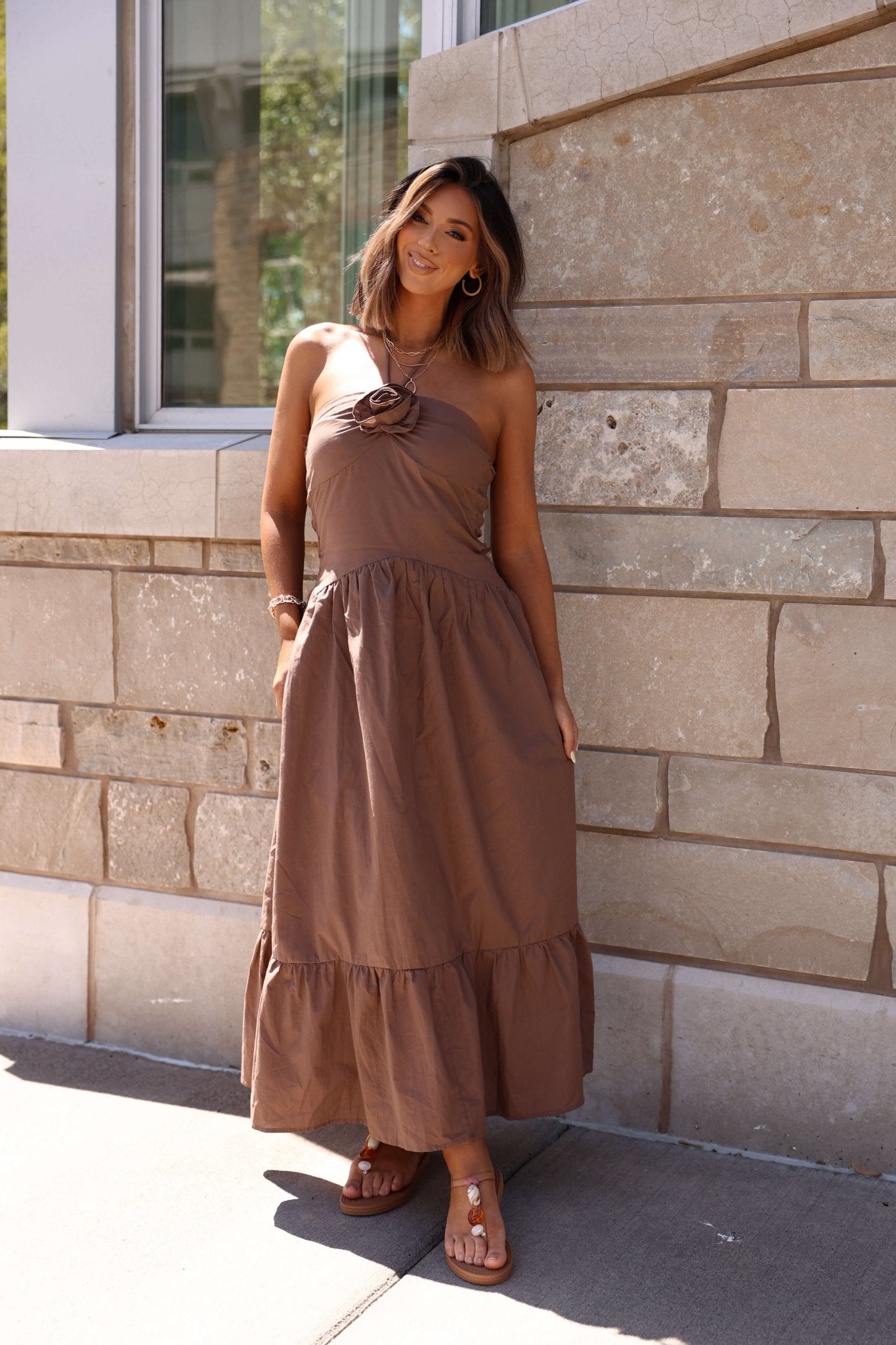 Woman in a Mocha Rosette Detail Halter Tiered Maxi Dress stands by a stone wall, smiling at the camera.