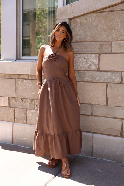 Woman in a Mocha Rosette Detail Halter Tiered Maxi Dress stands by a stone wall, smiling at the camera.
