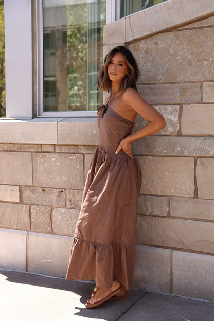 Woman poses against a stone wall in the Mocha Rosette Detail Halter Tiered Maxi Dress, standing near a large window.