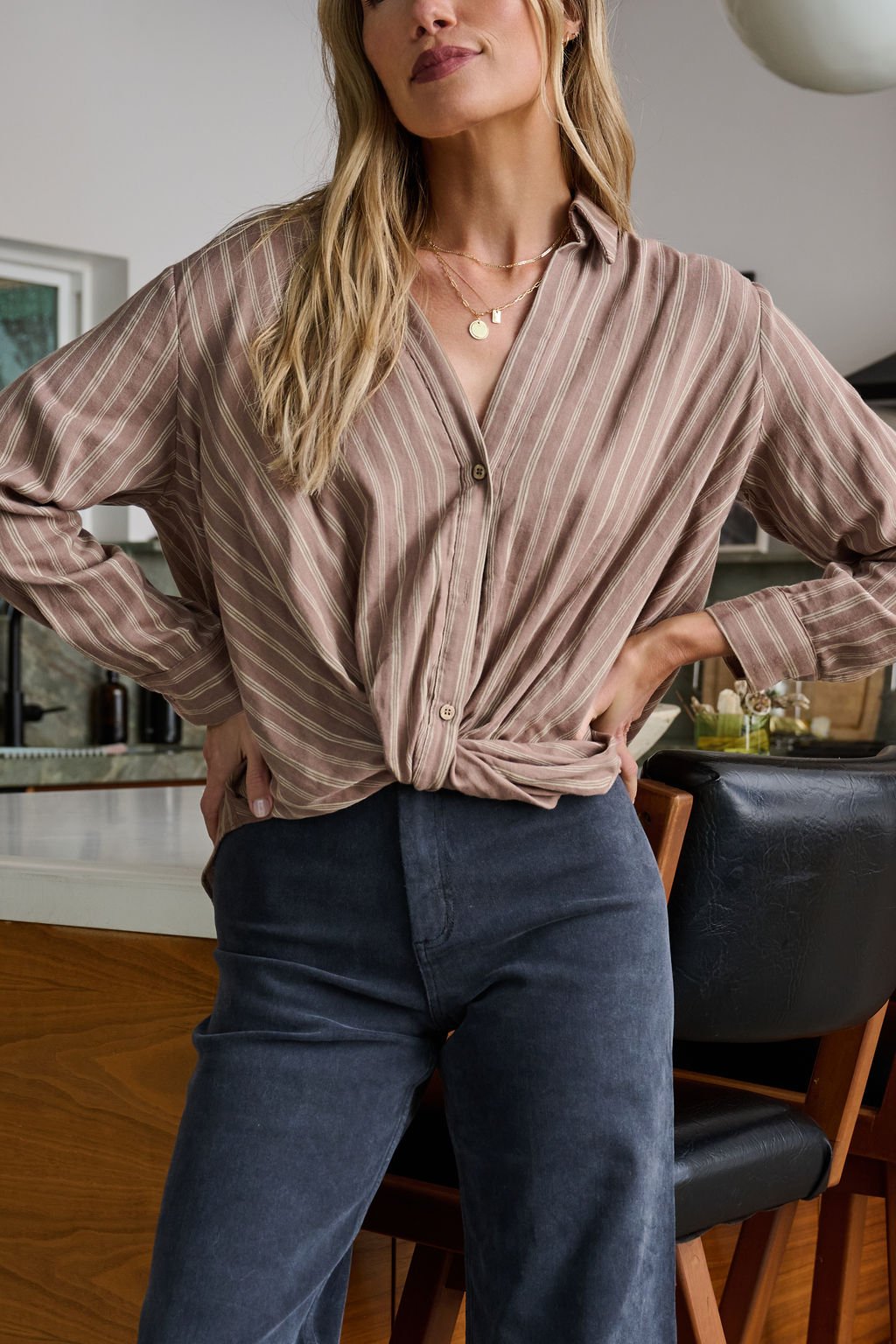 Woman stands confidently in a modern kitchen wearing the Mocha Striped Front Twist Top and dark pants.