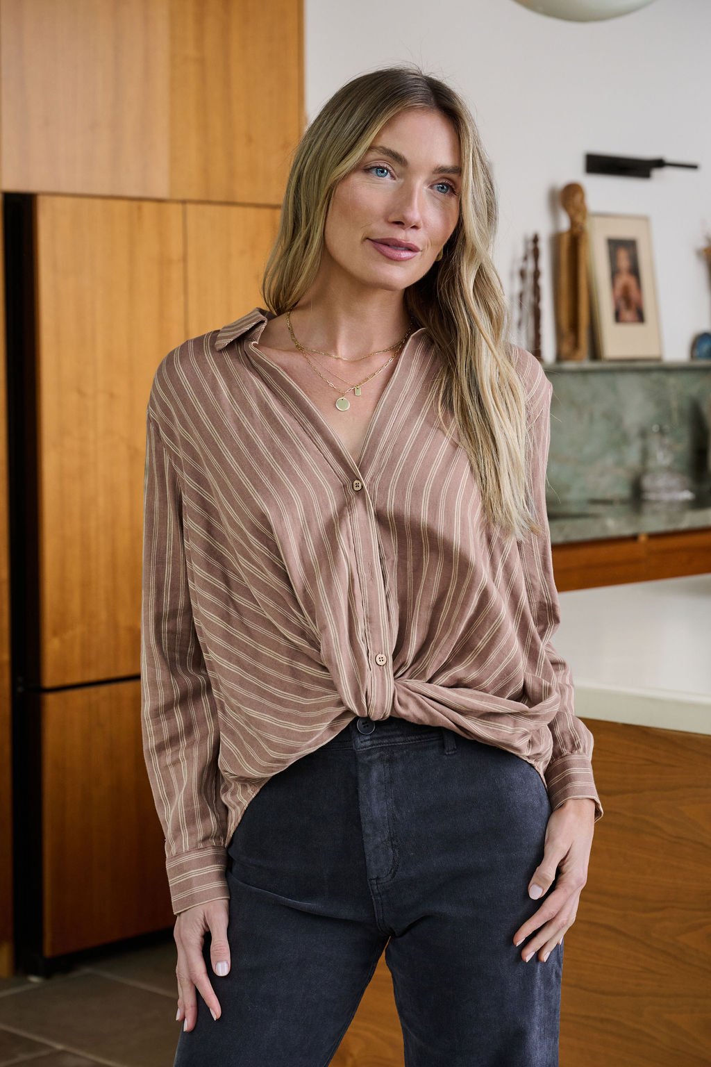 A woman in a Mocha Striped Front Twist Top with dark pants stands indoors, one hand in her pocket, looking to the side.