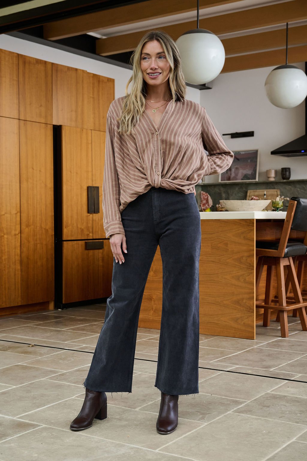 Wearing a Mocha Striped Front Twist Top and black wide-leg jeans, a woman smiles in a modern kitchen.