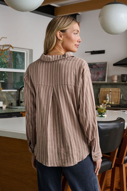 A woman stands in a kitchen, wearing the Mocha Striped Front Twist Top with a collared neckline and dark pants.