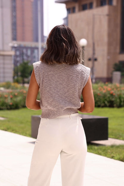 A woman with shoulder-length brown hair wears the Mocha Turtleneck Cap Sleeve Sweater and white pants, facing away outdoors.
