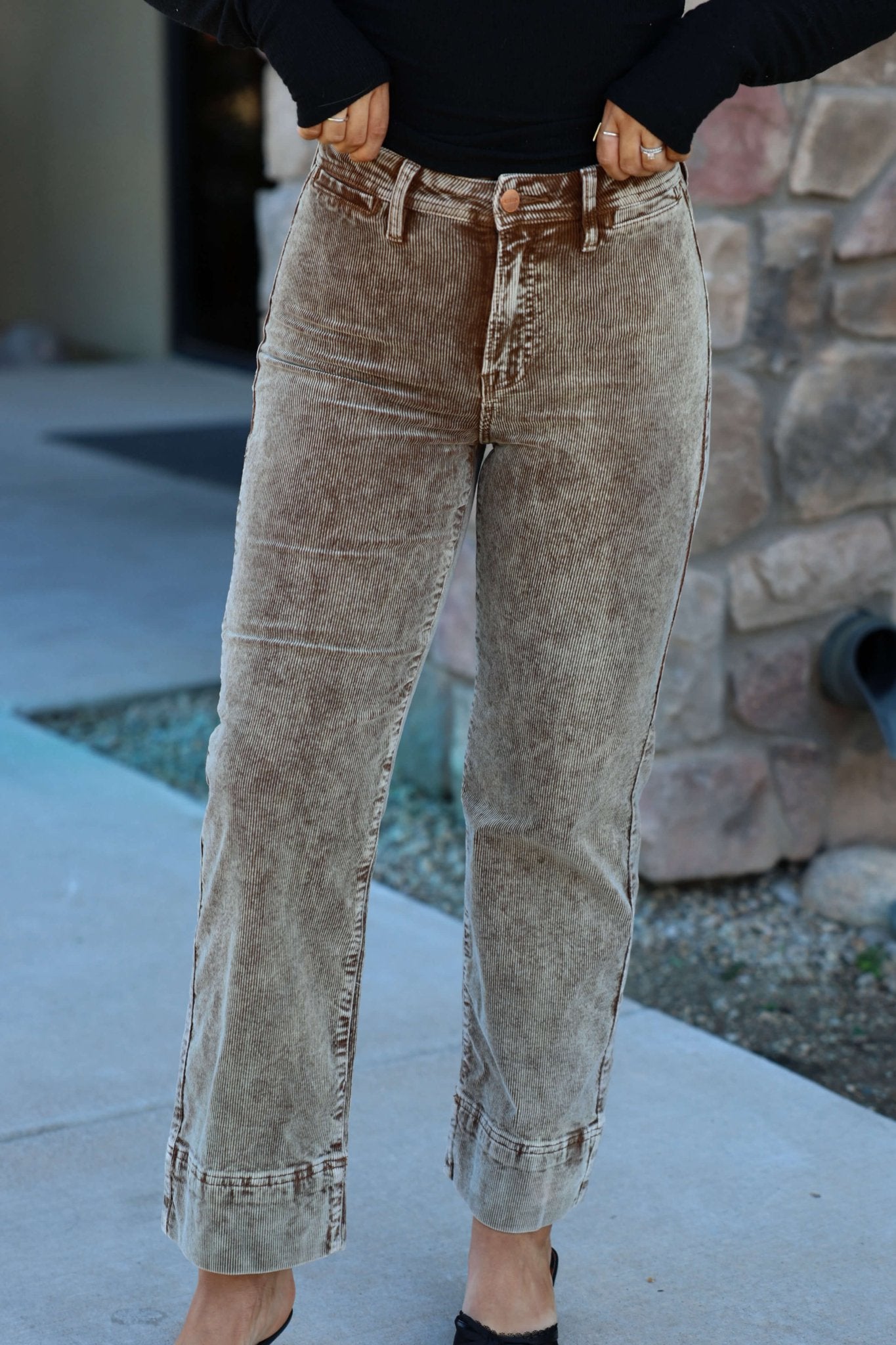 Person wearing Mocha Wash Wide Leg Cuffed Jeans and a black top, standing near a stone wall.