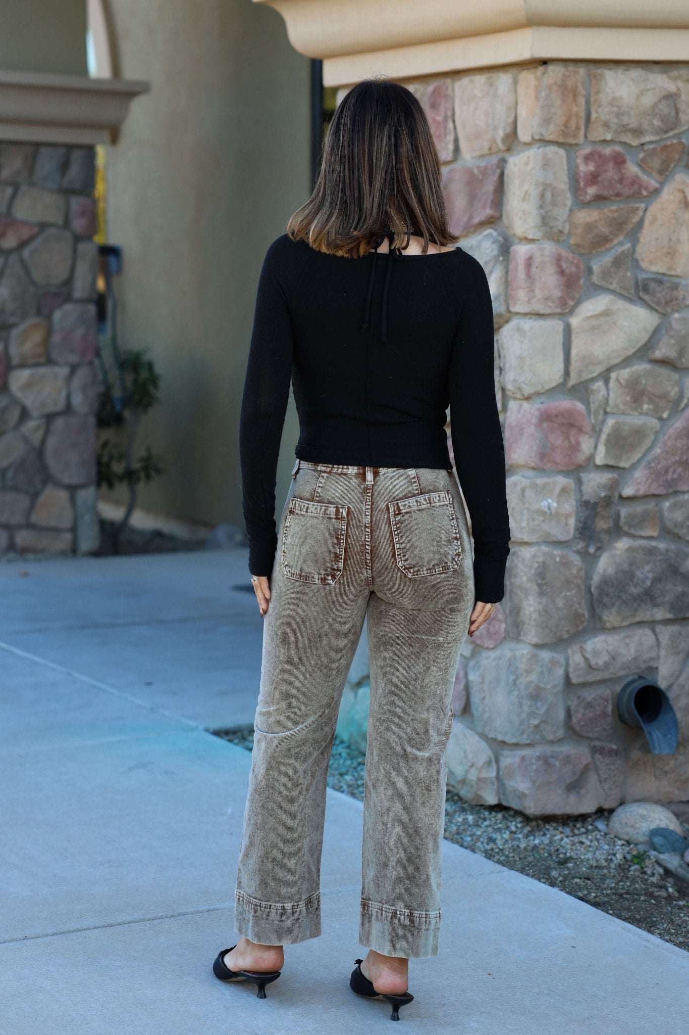 A woman with shoulder-length hair in a black top and Mocha Wash Wide Leg Cuffed Jeans stands outside by a stone building, facing away.
