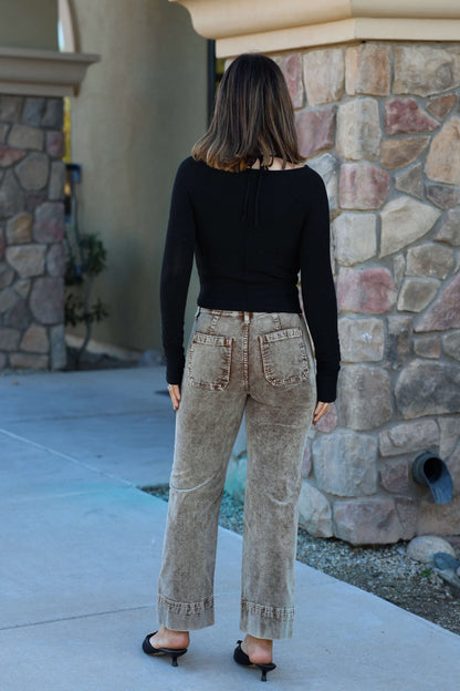 A woman with shoulder-length hair in a black top and Mocha Wash Wide Leg Cuffed Jeans stands outside by a stone building, facing away.