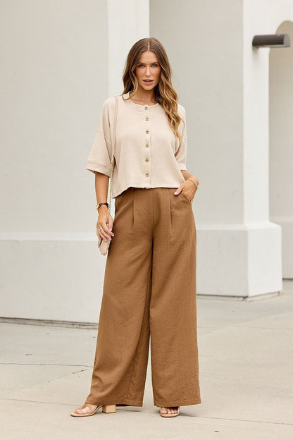 Woman wears the Monday Morning Button Down Top in oatmeal, styled with relaxed brown pants, in front of a classic white building.