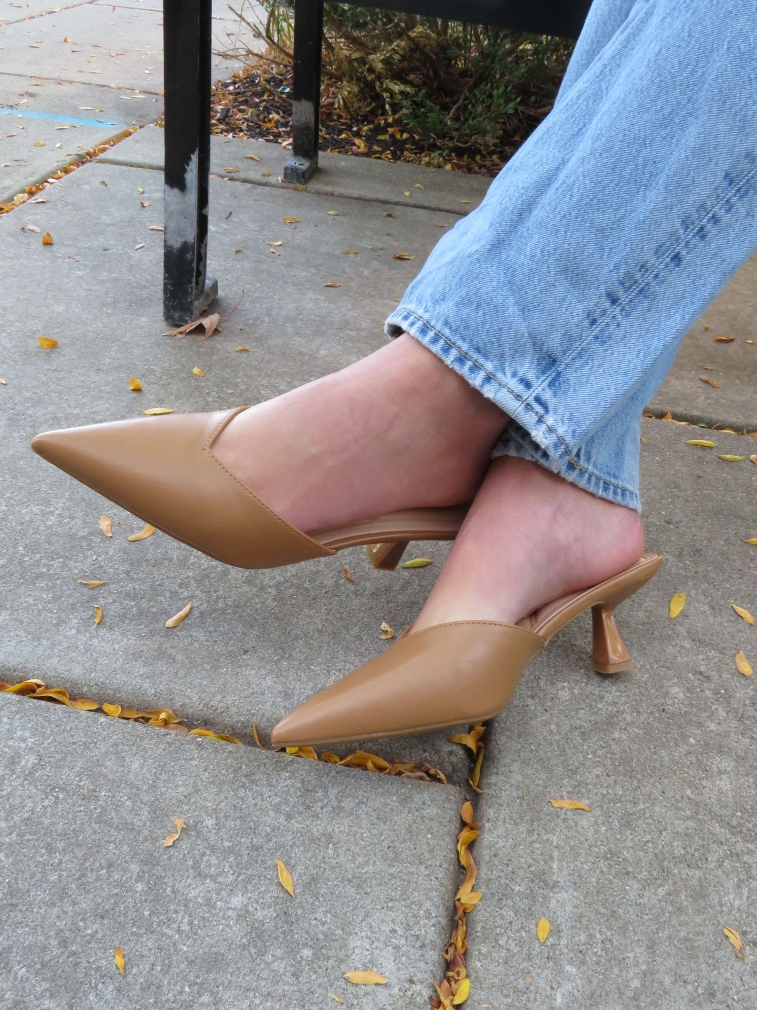 Person in light blue jeans and Morley Camel Open Back Kitten Heels sits on a bench, legs crossed amid scattered leaves.