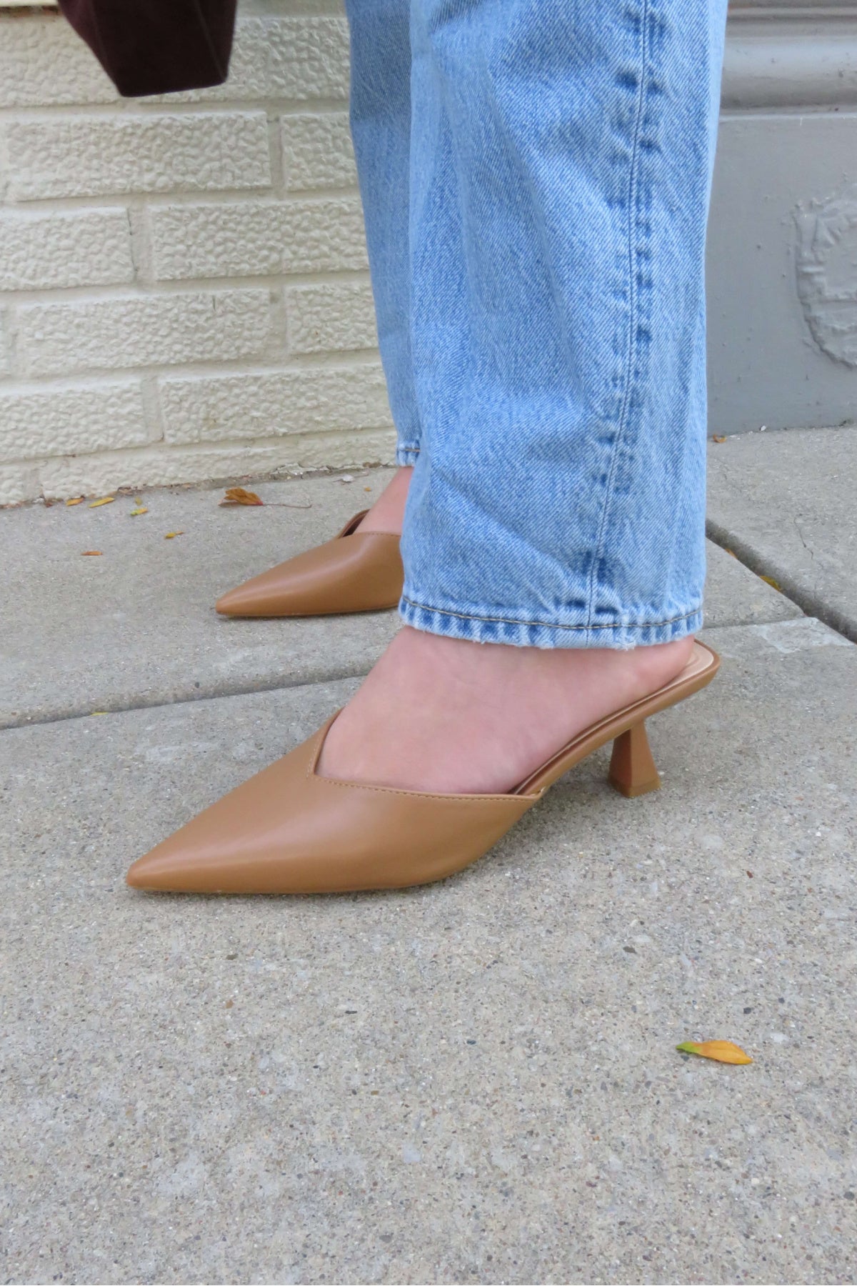 A person in light blue jeans and Morley Camel Open Back Kitten Heels stands on a sidewalk by a cream brick wall.
