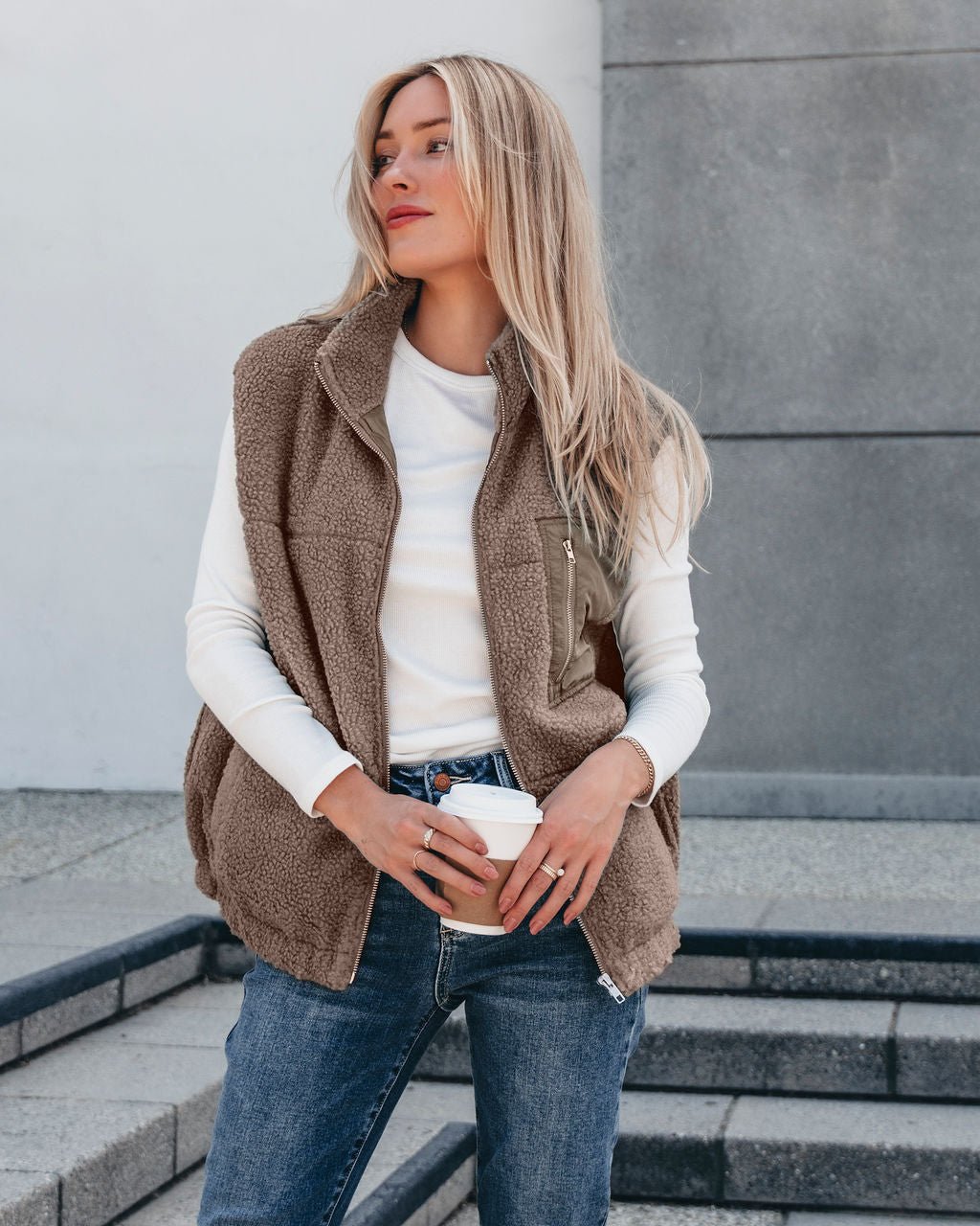 Woman in the Mountainside Zip Up Fleece Vest - Mocha, white shirt, and jeans holding coffee—perfect fall style on outdoor steps.
