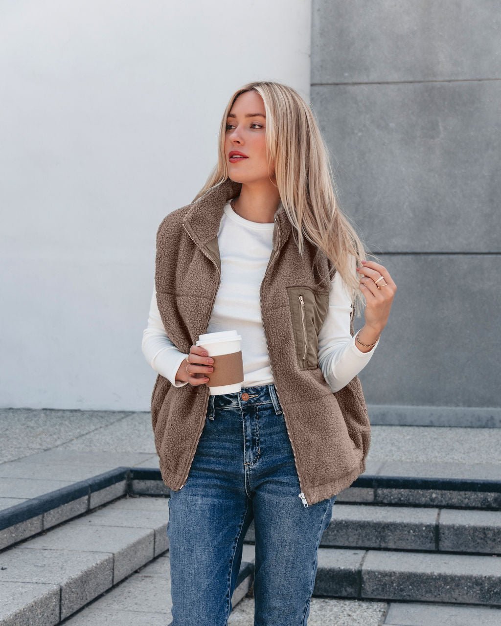 A woman wears the Mountainside Zip Up Fleece Vest in Mocha and jeans, holding a coffee cup on outdoor concrete steps.