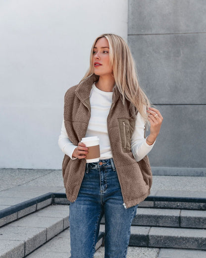 A woman wears the Mountainside Zip Up Fleece Vest in Mocha and jeans, holding a coffee cup on outdoor concrete steps.