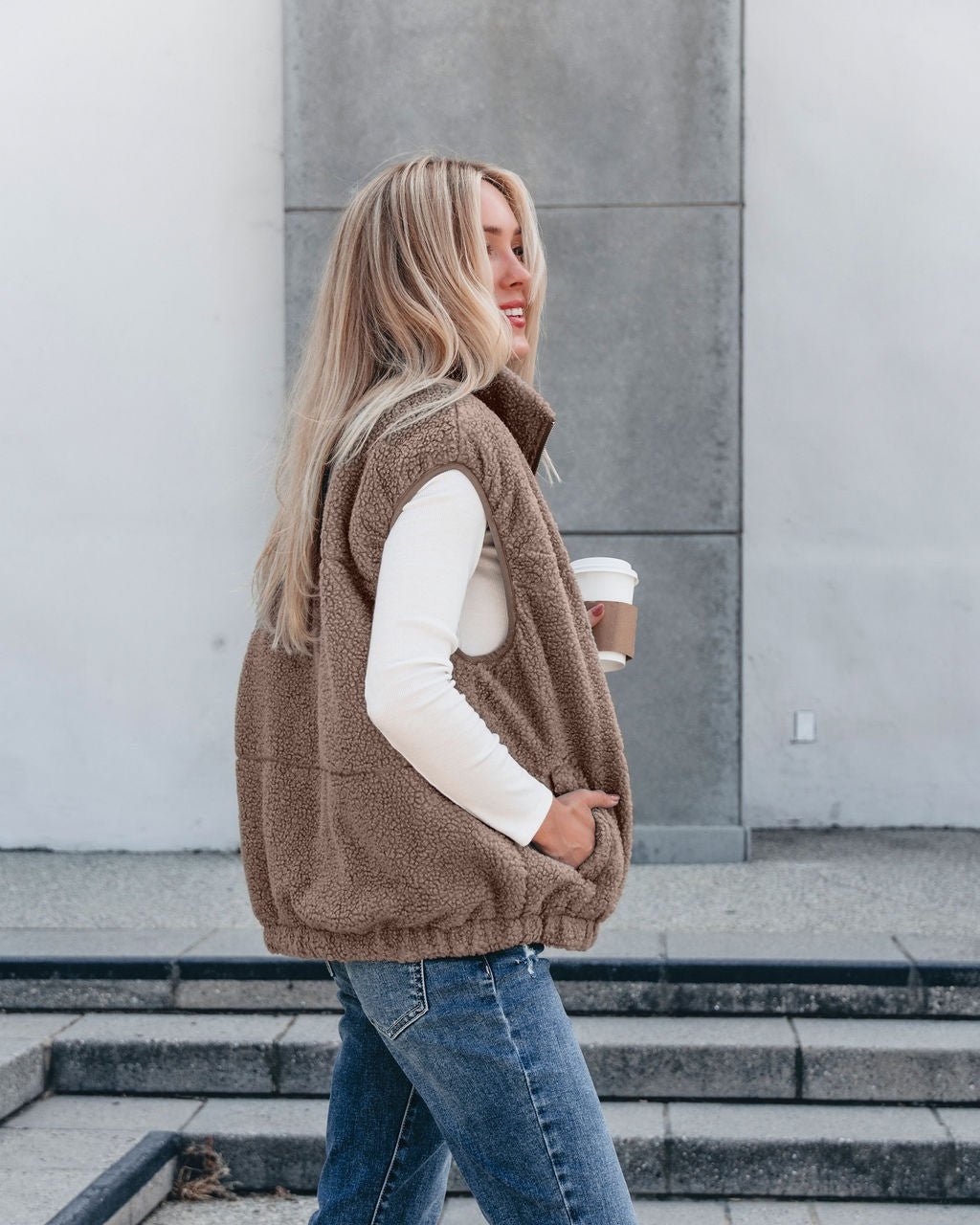Woman with long blonde hair wears the Mountainside Zip Up Fleece Vest in Mocha and jeans, holding coffee—cozy fall style.