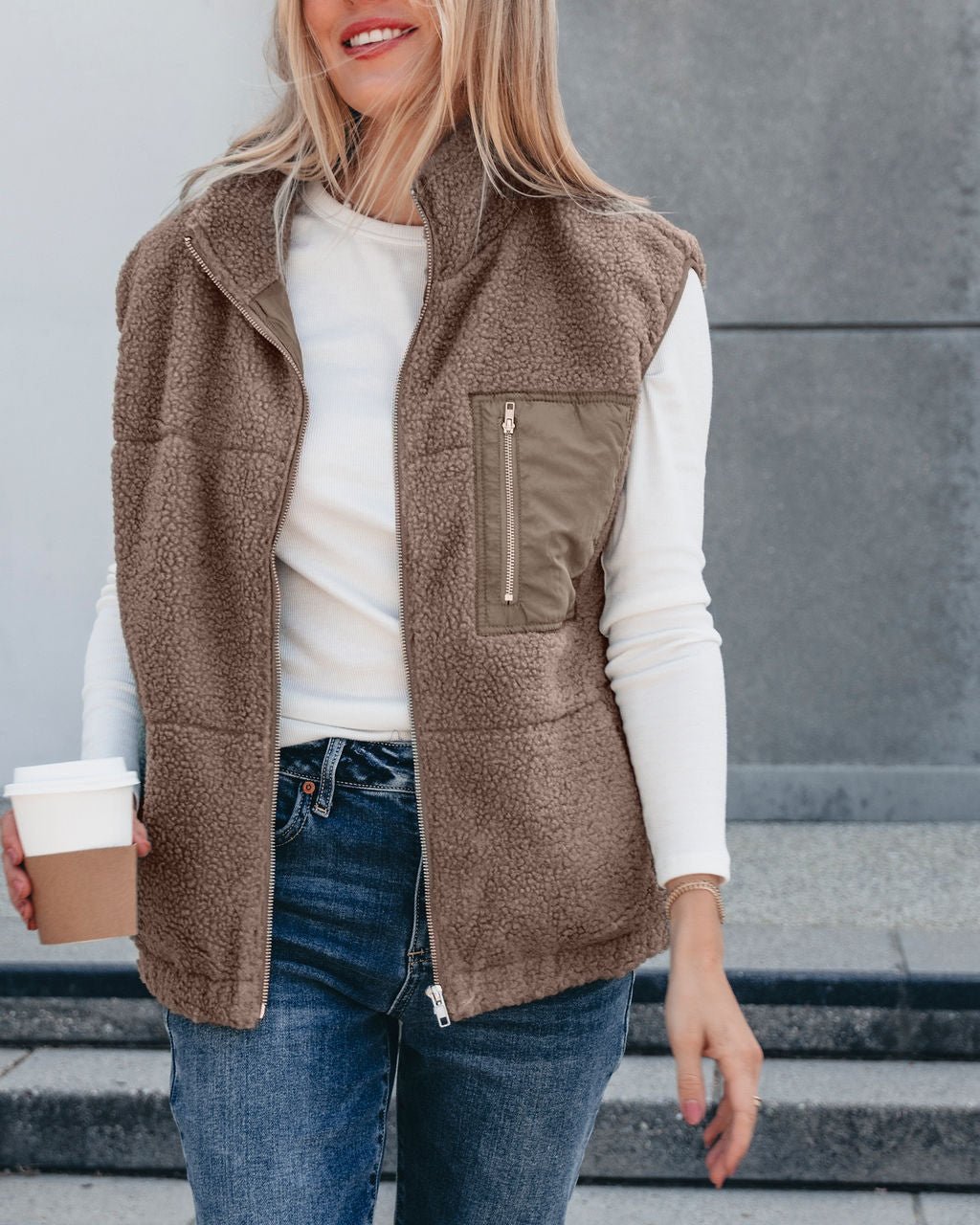 A woman wears the Mountainside Zip Up Fleece Vest - Mocha over a white shirt, holding coffee outdoors in a cozy fall look.