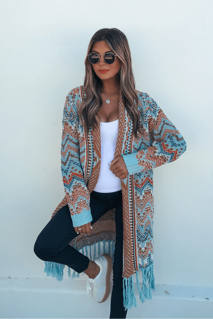 Woman wearing sunglasses, a Multi Aztec Stripe Boho Fringe Cardigan, black pants, and white shoes stands against a white wall.