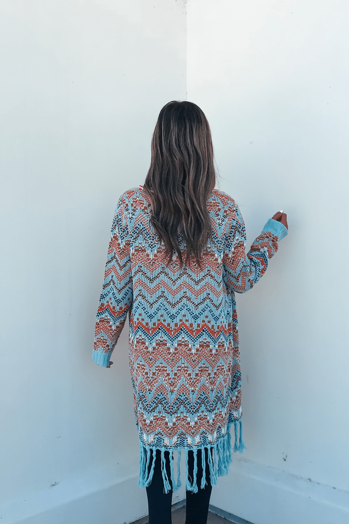 A woman with long hair wears a Multi Aztec Stripe Boho Fringe Cardigan, standing with her back to the camera facing a white wall.