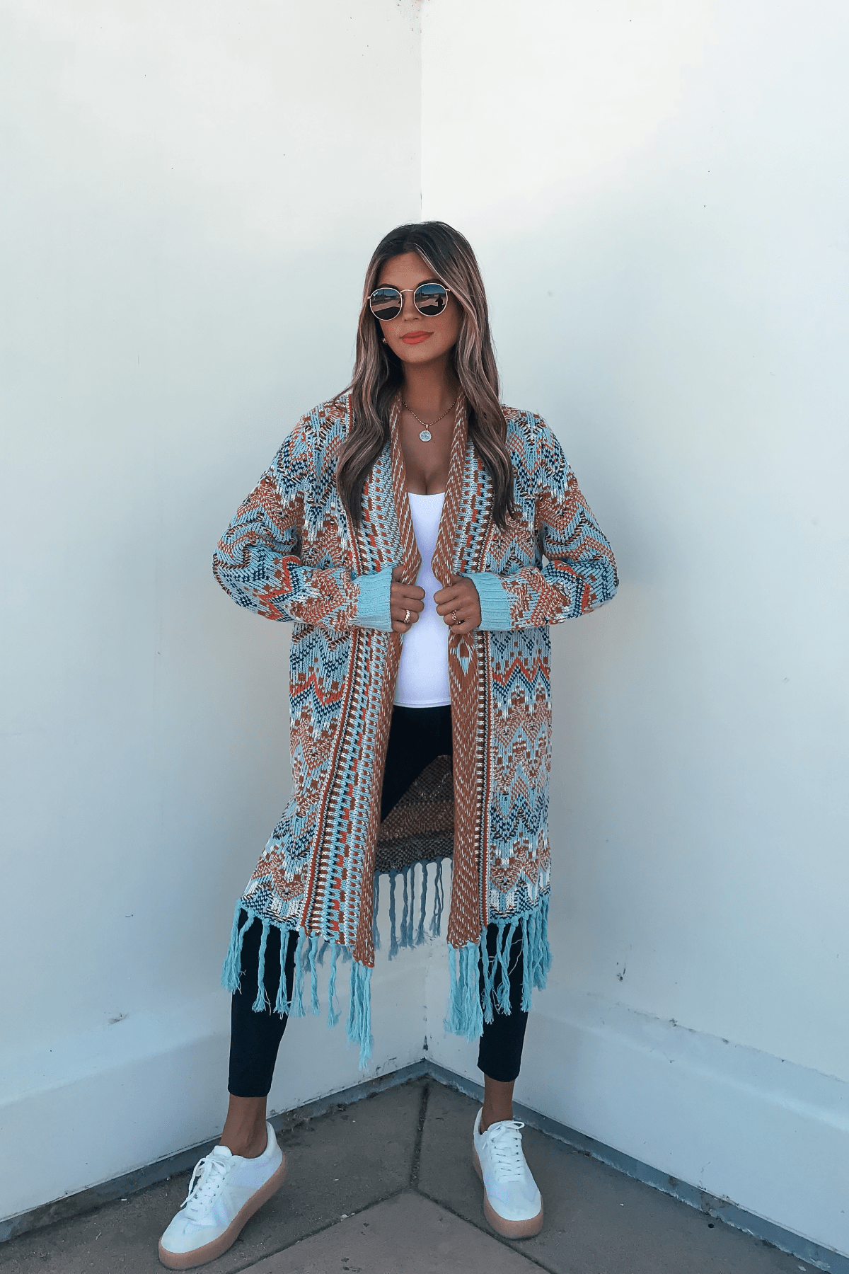 Woman in sunglasses and Multi Aztec Stripe Boho Fringe Cardigan with black leggings, white sneakers, and top stands by a white wall.