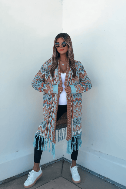 Woman in sunglasses and Multi Aztec Stripe Boho Fringe Cardigan with black leggings, white sneakers, and top stands by a white wall.