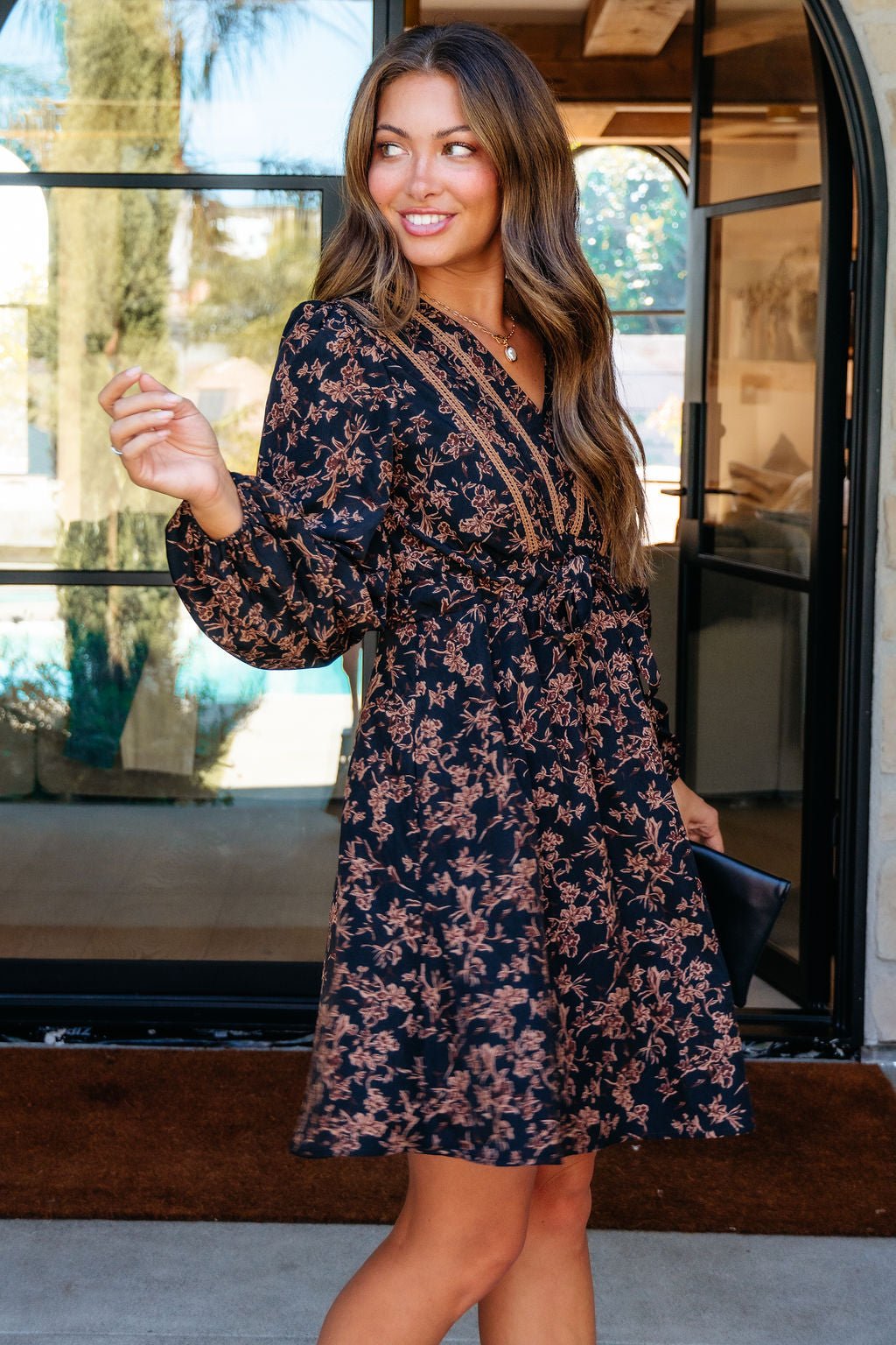 Woman smiles in front of glass doors, wearing the Multi Black Floral Print Mini Dress and holding a black clutch.
