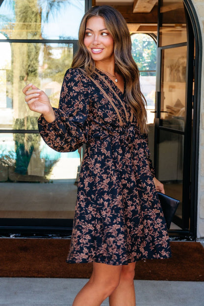 Woman smiles in front of glass doors, wearing the Multi Black Floral Print Mini Dress and holding a black clutch.
