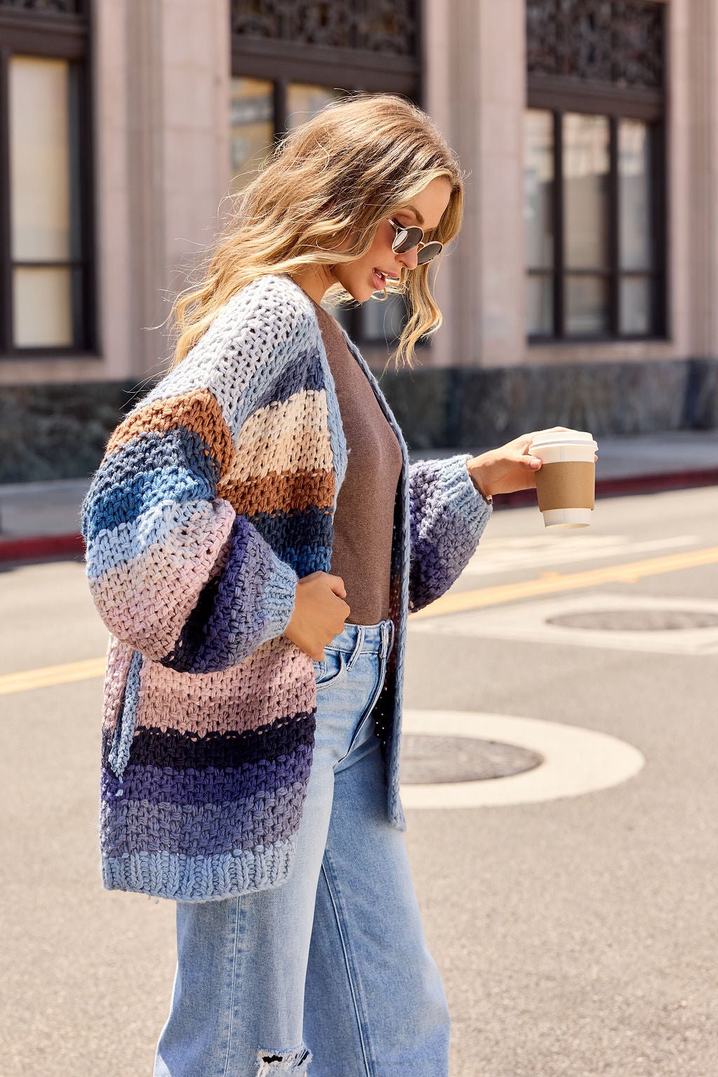 Woman in a Multi Blue Stripe Crochet Cardigan, sunglasses, and jeans walks outside holding coffee on a city street.